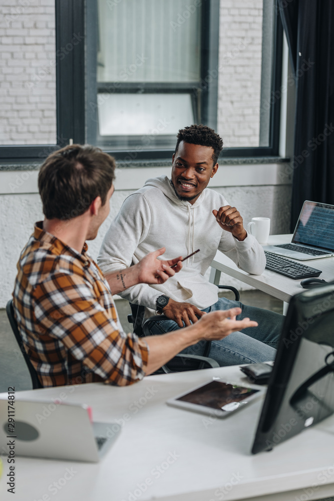 two cheerful multicultural programmers discussing ideas while sitting together in office two cheerful multicultural programmers discussing ideas while sitting together in office