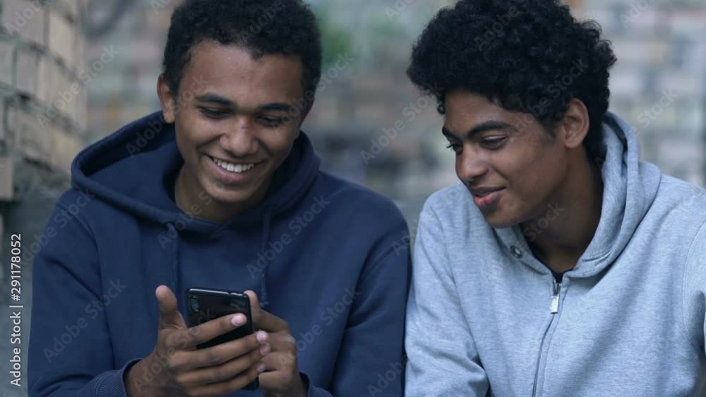 Black male teenager showing friend photos on smartphone, having fun outdoors Black male teenager showing friend photos on smartphone, having fun outdoors