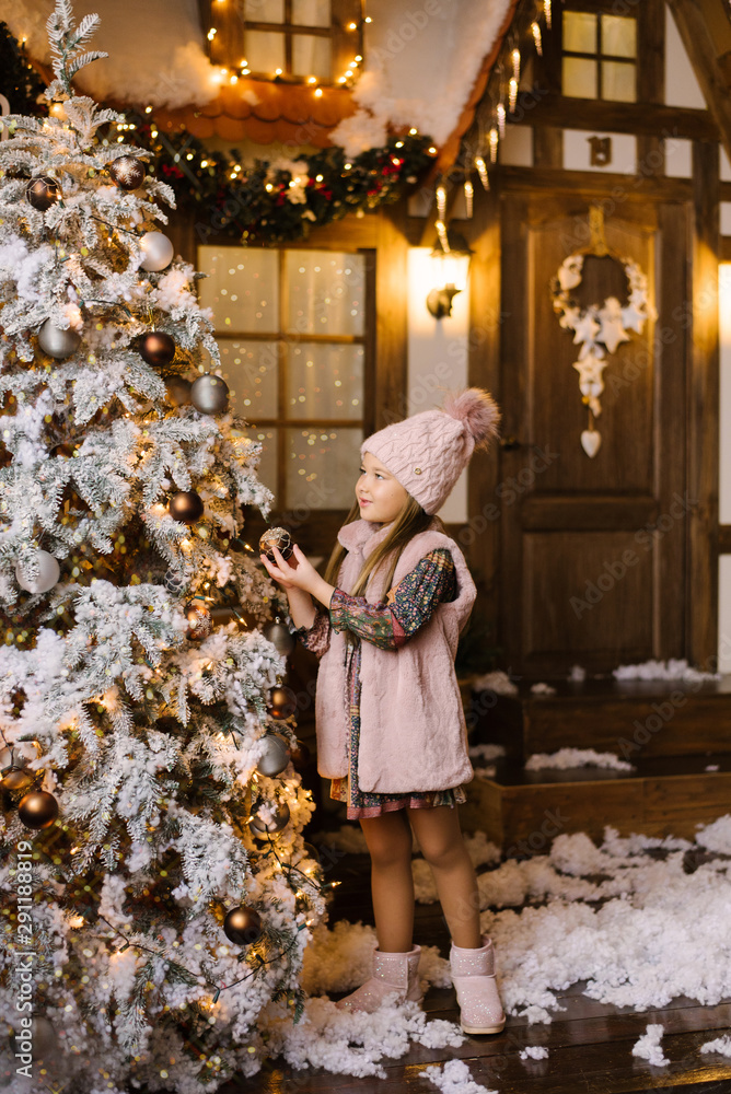 A girl of five or six years in winter pink clothes and ugg boots stands near the Christmas tree and decorates it with balls on the background of a winter house. Christmas story