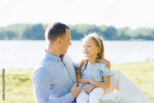 A little girl four or five years old sits on dad's lap against the lake in the summer, they look at each other and smile © Sunshine