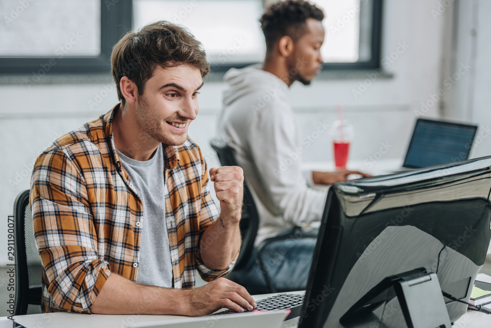 cheerful programmer showing winner gesture while working near african american colleague cheerful programmer showing winner gesture while working near african american colleague
