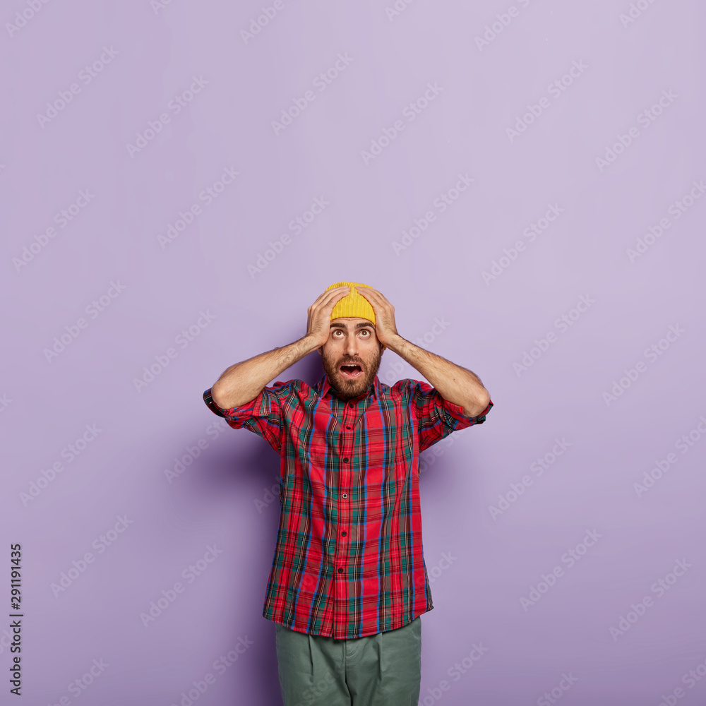 Photo of surprised emotional guy keeps both hands on head, wears yellow hat and checkered shirt, has atonished look upwards, feels puzzled and frustrated, isolated on purple background, being in panic Photo of surprised emotional guy keeps both hands on head, wears yellow hat and checkered shirt, has atonished look upwards, feels puzzled and frustrated, isolated on purple background, being in panic