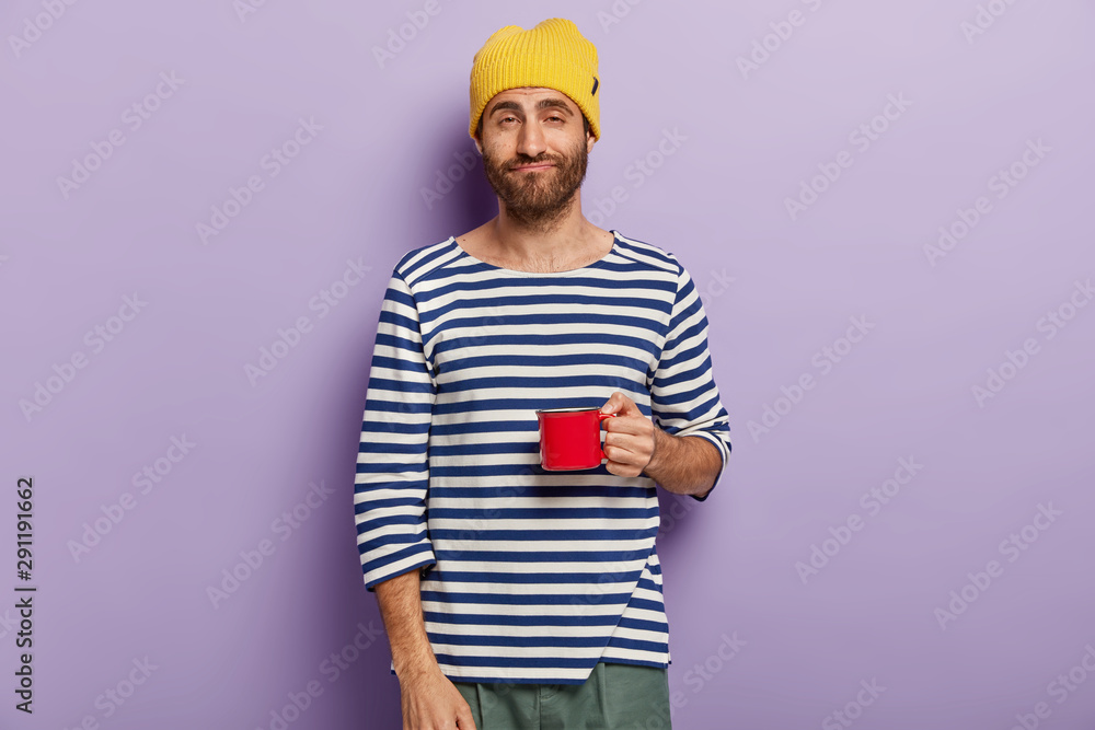 Photo of sleepy dissatisfied man has morning routine, being tired after sleepless night, wears yellow hat and striped jumper, isolated over purple background. Caucasian guy holds mug of fresh drink
