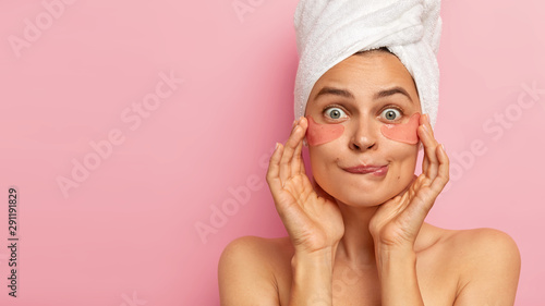 Cropped image of lovely woman bites lips, applies collagen patches under eyes, looks surprisingly at camera, touches cheeks with both hands, wears wrapped towel, stands against pink wall, copy space