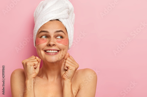 Indoor shot of happy European woman clenches teeth and fists, looks away with glad expression, wears white soft towel on head, has naked shoulders, stands over pink wall, empty space for promo