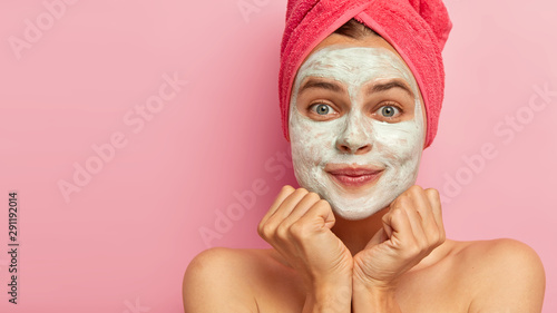 Cropped shot of pleased young European female applies facial clay mask, has beauty treatments, cares about complexion and body, stands against rosy studio wall, copy space for your advertisement