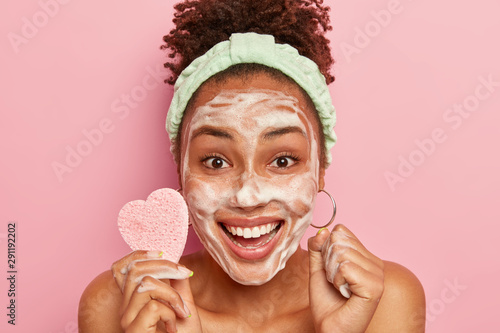 Cropped image of happy African American woman enjoys relaxing time, washes face with soap bubble, feels refreshed and delighted, holds cosmetic sponge for wiping complexion, isolated on pink wall