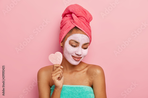 Принти на полотні Pleased young dark skinned woman focused down, applies facial clay mask, holds cosmetic sponge for removing makeup, shows bare shoulders, wrapped in bath towel, isolated on pink studio wall