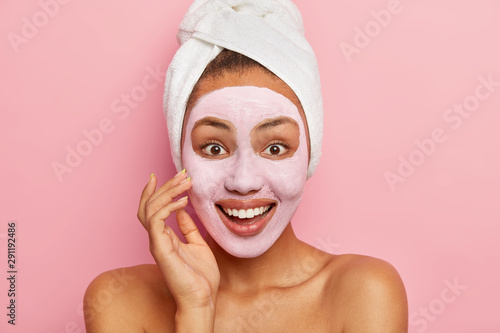 Close up portrait of lovely happy young woman applies facial mask on complexion, uses nourishing clay for rejuvenation, wears wrapped white towel on head, stands naked against pink background.