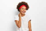 Happy dark skinned young woman with curly hair, touches neck, enjoys pleasant moment in life, dressed in casual clothes, isolated on white background, being in good mood, wears silver earrings