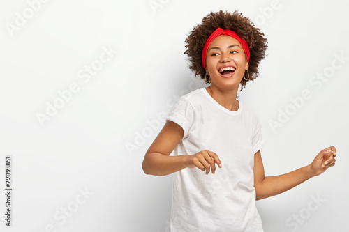 Photo of pretty dark skinned woman stands sideways, feels energized dances actively to music wears red headband casual white t shirt poses indoor. Horizontal shot of lovely African American girl moves