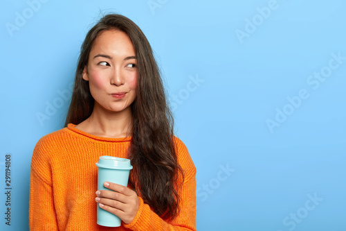Photo of pensive brunette woman purses lips, looks thoughtfully aside, holds takeout coffee, makes decision in mind, plans her day, wears orange jumper, stands over blue wall. Asian girl with beverage