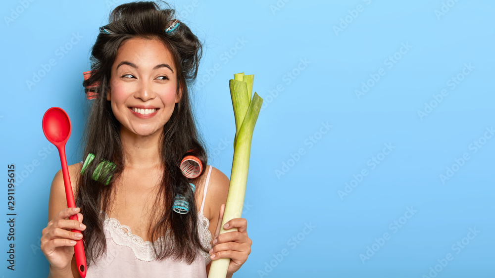 Positive young Korean woman with gentle smile, holds red spoon and green leek, busy preparing healthy breakfast, keeps to diet for being fit, wears curlers and nightgown, isolated over blue wall Positive young Korean woman with gentle smile, holds red spoon and green leek, busy preparing healthy breakfast, keeps to diet for being fit, wears curlers and nightgown, isolated over blue wall