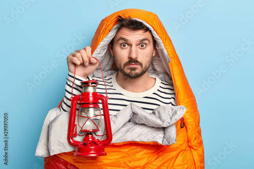 Surprised man has rest, wrapped in sleeping bag, holds kerosene lamp, enjoys recreation in forest, has expedition, poses against blue background, has adventure trip. People, lifestyle and rest concept photo