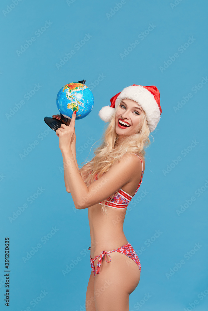 Happy woman wearing Santa hat and bikini selects a tropical destination during winter holidays on the globe. Blue isolated background. Christmas vacation and travel on the beach