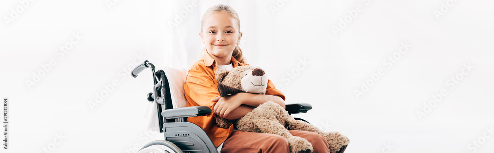 panoramic shot of smiling kid holding teddy bear and sitting on wheelchair - obrazy, fototapety, plakaty 