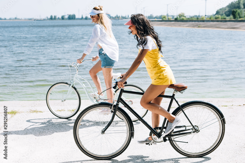 Fototapeta premium side view of smiling blonde and brunette girls riding bikes near river in summer