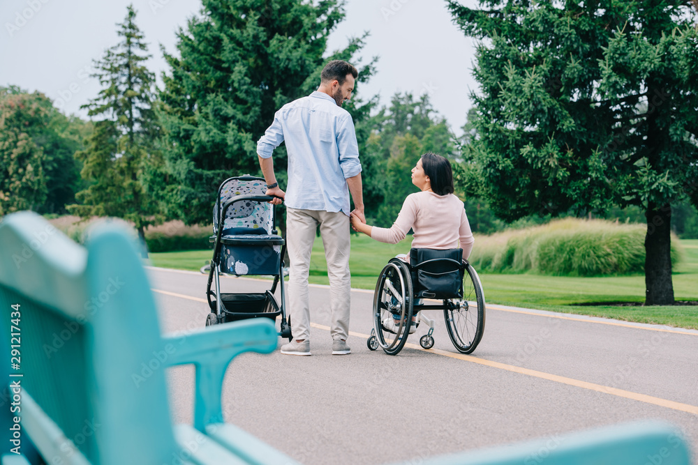 selective focus of happy disabled woman holding hands with husband while walking with baby carriage in park together