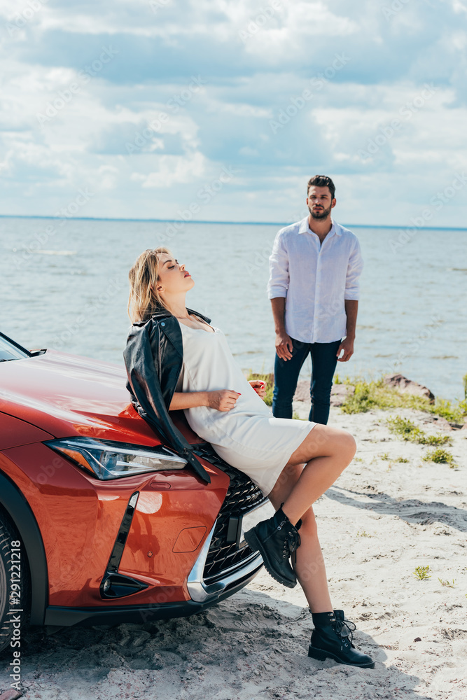 attractive woman in dress with closed eyes lying on car and man look at her