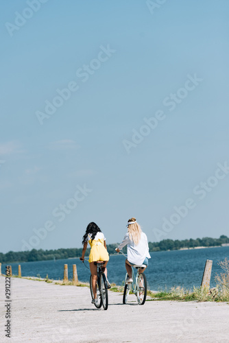 back view of blonde and brunette friends riding bikes near river in summer