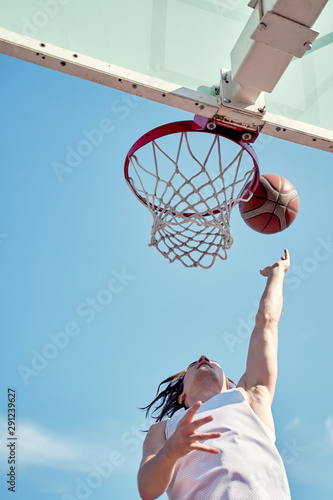 Photo of athlete man throwing ball into basketball hoop on sports field on street on summer day.