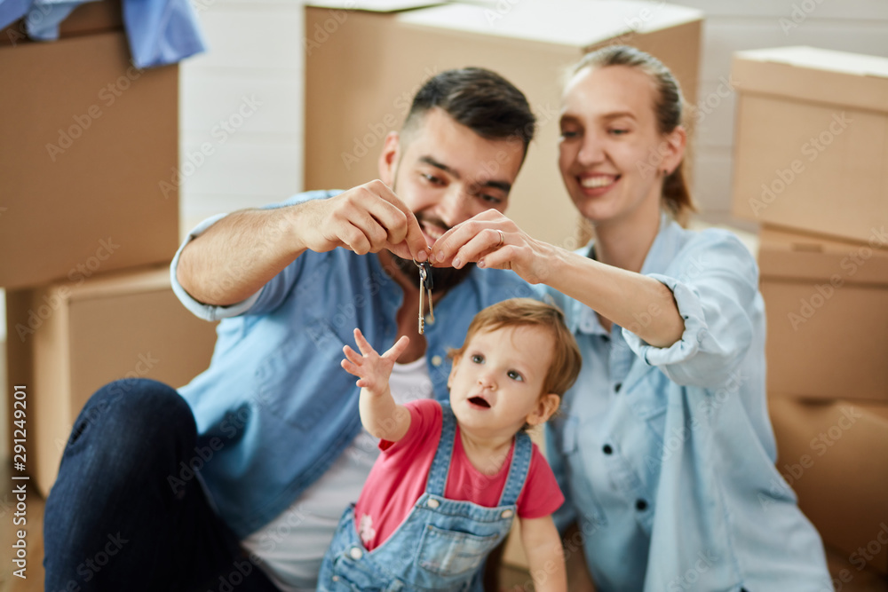 Family sit on floor near open box. Man and woman hold keys. Camera focus keys. Background moving boxes Family sit on floor near open box. Man and woman hold keys. Camera focus keys. Background moving boxes