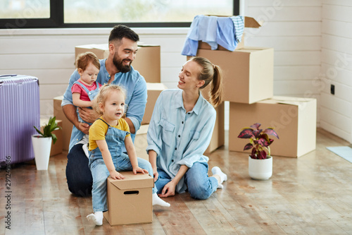 Family of four men sit on floor in guest room in just sell house. Eldest daughter sit on box like rider. Mom and dad smile. Dad keep youngest daughter. Background moving boxes, suit and flower in pot