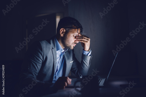 Tired hardworking handsome Caucasian businessman sitting in office late at night and rubbing eyes. In front of his is laptop. Overworking concept. photo