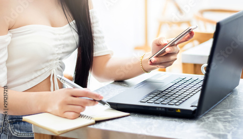 Closeup of hand businesswoman using smartphone and laptop working on desk and writing on notebook