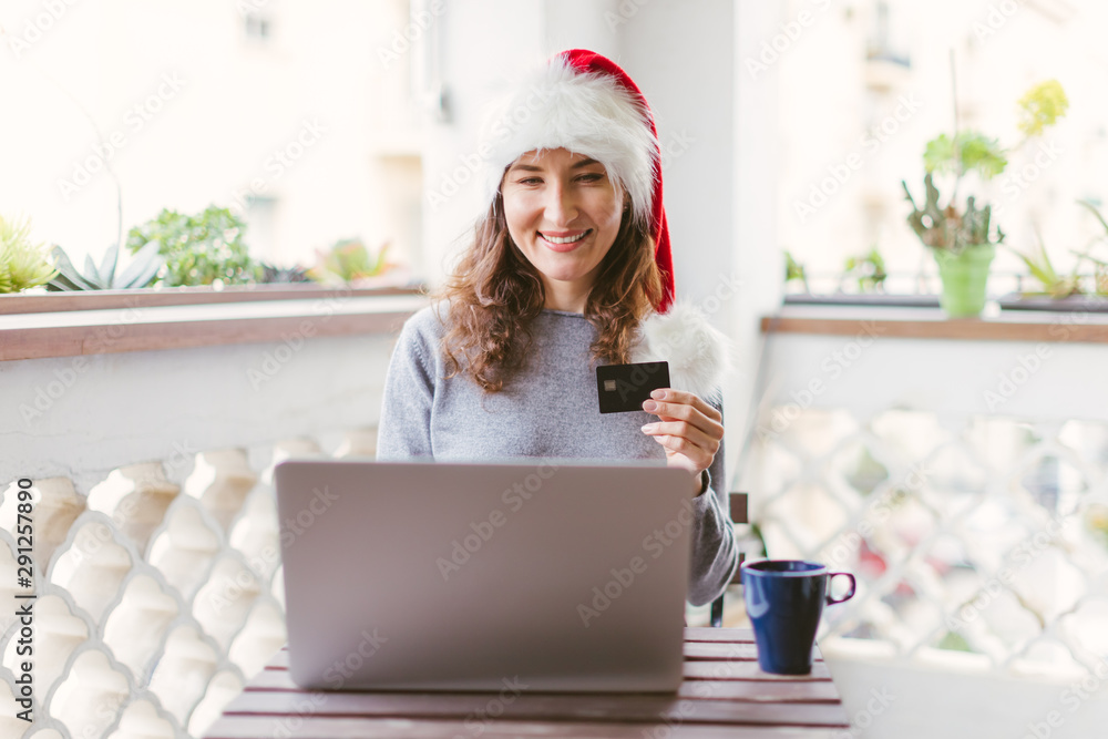 Happy smiling woman sitting at the table with laptop and credit card wearing Santa's red hat - Christmas and new year holiday gift shopping online, winter sale and spending money concept Happy smiling woman sitting at the table with laptop and credit card wearing Santa's red hat - Christmas and new year holiday gift shopping online, winter sale and spending money concept