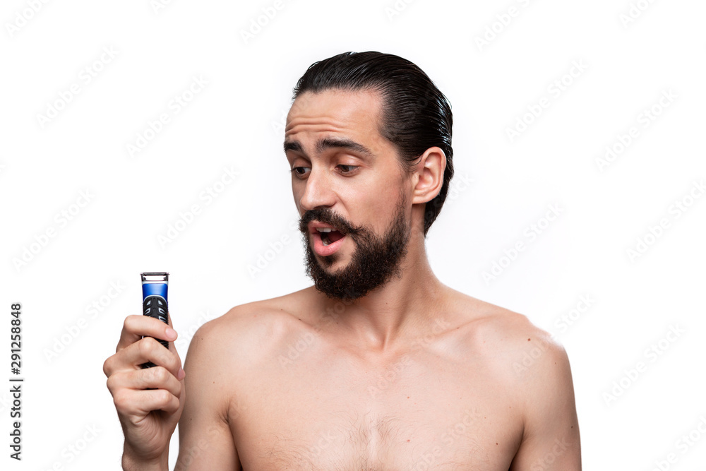Fototapeta premium Handsome bearded man with mustache holds electric shaver standning bare isolated over white background. Concept of morning treatment and shaving. Time to trim your beard. Morning routine