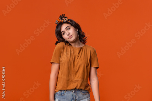 Unsure brunette woman in a t-shirt and beautiful headband shrugging shoulders as if she doesn't know the answer standing isolated over orange background. Place for ad. © ianachyrva