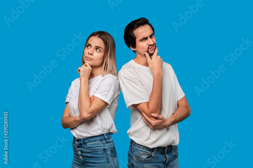 Thoughtful couple of a bearded brunet man with mustache and blond woman in white t-shirts and blue jeands standing isolated over white background.