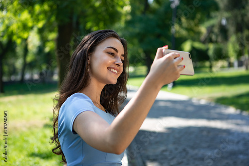 Smiling young woman in a blue dress sits in a park and making selfie with the smartphone.