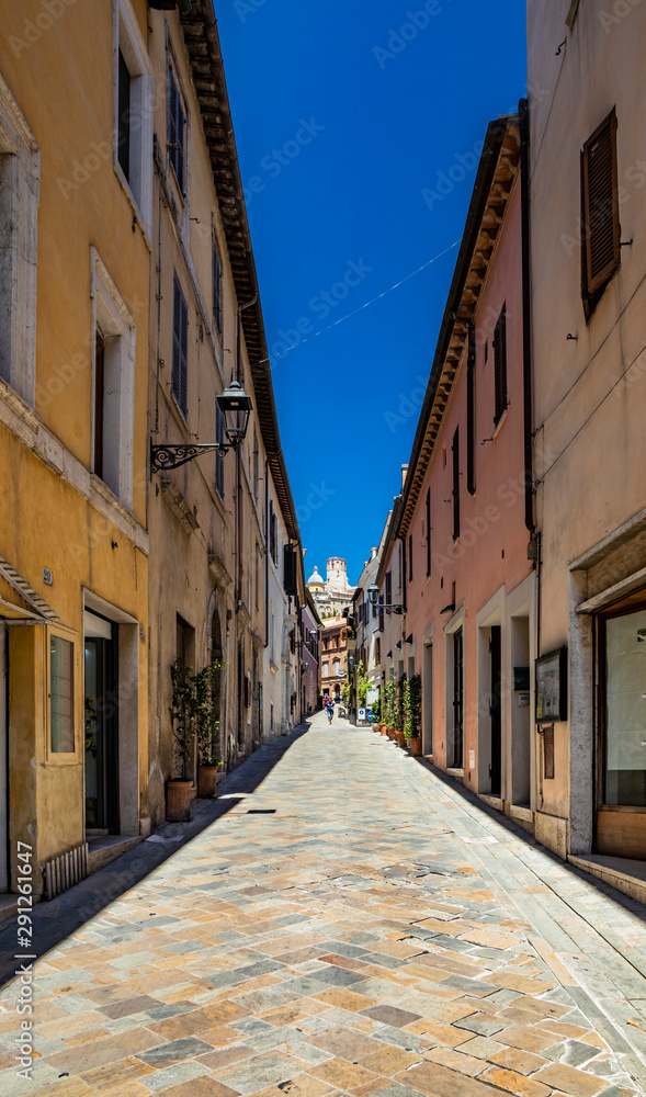 A narrow alley in the city of Amelia, in Umbria. The windows with closed doors and flowered balconies. The straight road in perspective leads towards the center of the village.