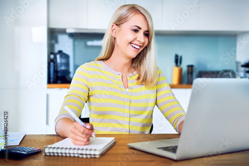 Smiling young woman sitting at home with laptop and notebook managing personal finance