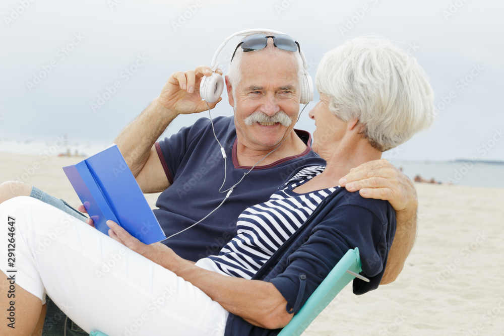 senior couple on beach reading book and listening to headphones