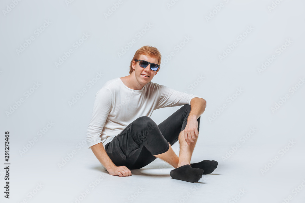 A red-haired man in sunglasses and a white t-shirt and black shorts sits on the floor against a light background A red-haired man in sunglasses and a white t-shirt and black shorts sits on the floor against a light background