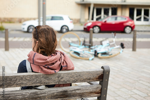 Rear view of young female with sunglasses on head and scarf around neck sitting on wooden bench with back to camera, relaxing while sightseeing in European town, bicycle on sidewalk in background