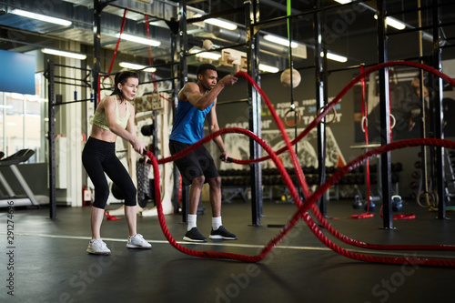 Side view portrait of fit couple exercising with battle ropes during strength workout in modern gym, copy space