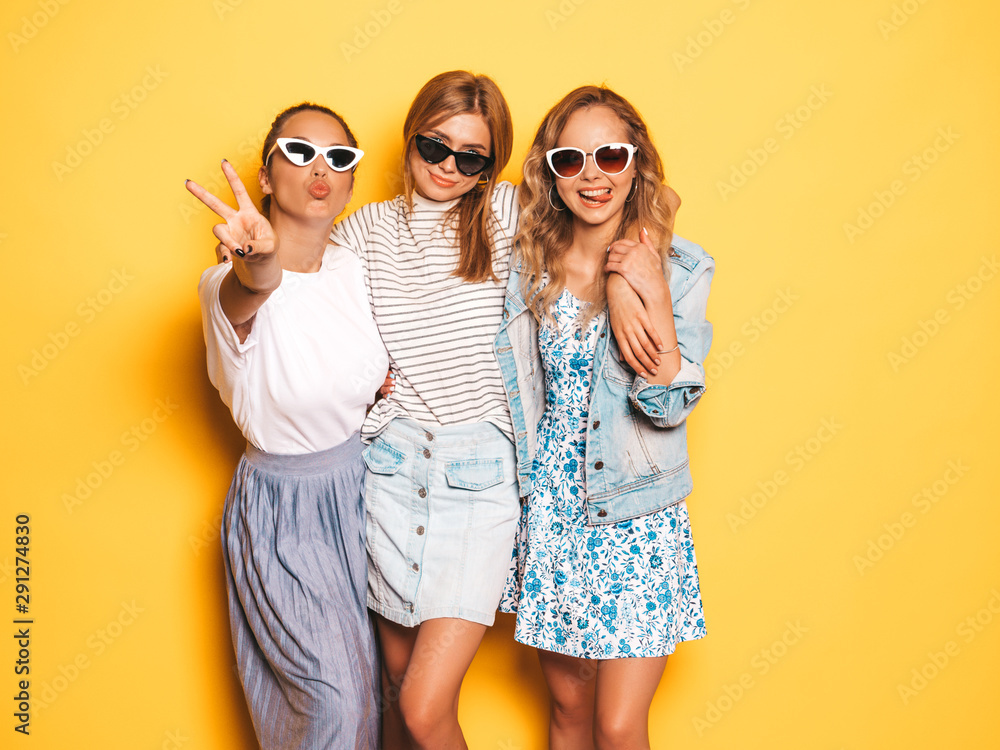 Three young beautiful smiling hipster girls in trendy summer clothes. Sexy carefree women posing near yellow wall in studio. Positive models going crazy and having fun in sunglasses - obrazy, fototapety, plakaty 