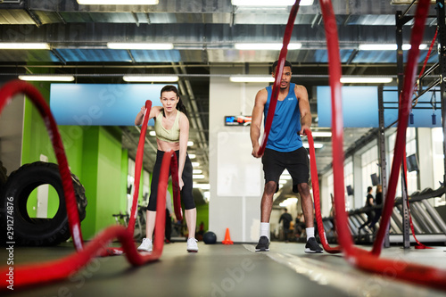 Front view portrait of fit couple exercising with battle ropes during strength workout in modern gym, copy space