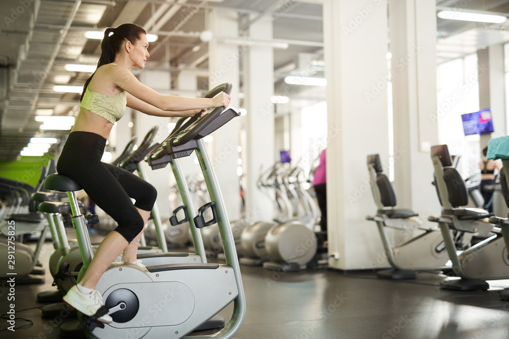 Side view portrait of fit young woman on exercise bike enjoying cardio workout alone in empty gym, copy space Side view portrait of fit young woman on exercise bike enjoying cardio workout alone in empty gym, copy space