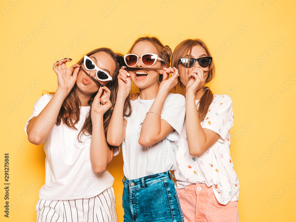 Three young beautiful smiling hipster girls in trendy summer clothes. Sexy carefree women posing near yellow wall in studio. Positive models going crazy and having fun.Making mustache with hair