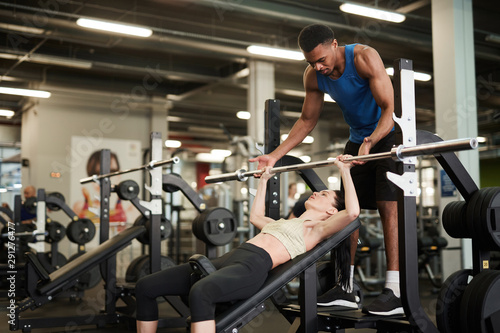 Portrait of African-American fitness coach helping young woman lifting weights on bench in modern gym, copy space