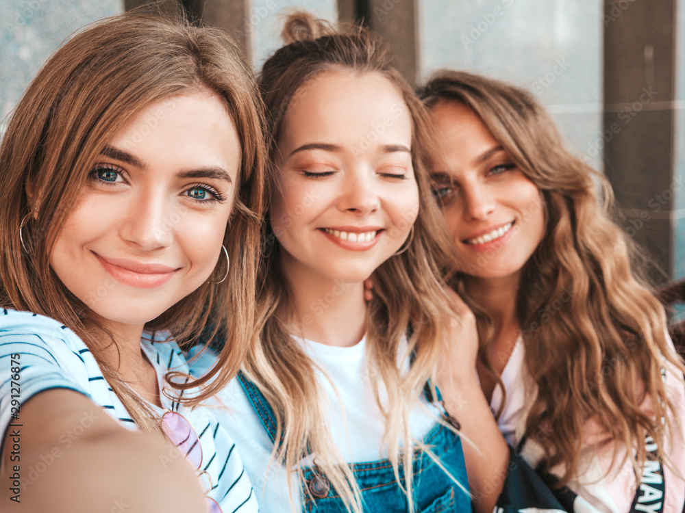 Three young smiling hipster women in summer clothes.Girls taking selfie self portrait photos on smartphone.Models posing in the street.Female showing positive face emotions