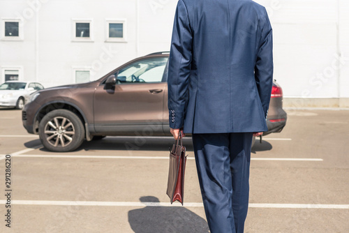 Middle-aged tall gray-haired businessman in blue suit with brown briefcase walks to his car on the private parking