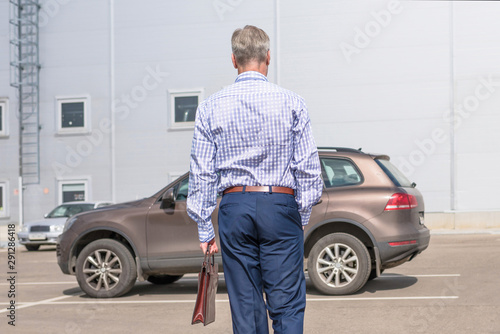 Middle-aged  tall gray-haired businessman in blue suit with brown briefcase walks to his car on the private parking © flowertiare