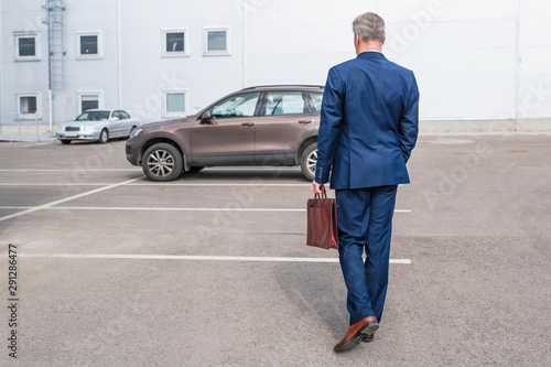 Middle-aged tall gray-haired businessman in blue suit with brown briefcase walks to his car on the private parking