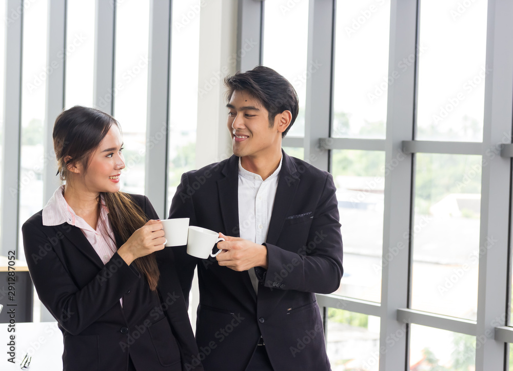 Obraz premium Couple of happy Asian colleagues talking while standing at the office window with cups of coffee.Concepts about teamwork, work fun and love
